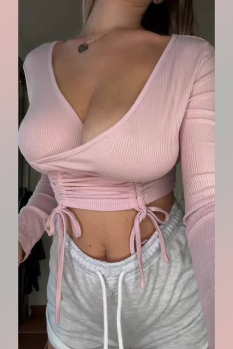 Close-up of a busty social media model with long hair and natural breasts performing hot moves in a pink top and grey shorts.