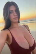 Beautiful busty brunette influencer in a plunging hot outfit showing huge cleavage at sunset on the beach, high-quality NSFW social media reveal.