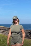 Gorgeous plus-size independent creator with an hourglass frame posing outdoors against a green landscape, wearing form-fitting tight pants and a thin braless top.