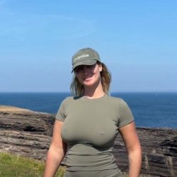 Gorgeous plus-size independent creator with an hourglass frame posing outdoors against a green landscape, wearing form-fitting tight pants and a thin braless top.