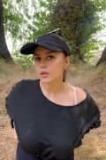 Cute independent creator holding her phone for an outdoor vertical video vlog, smiling warmly while walking along a scenic nature trail under natural sunlight.