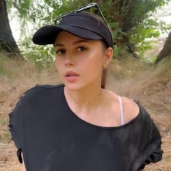 Cute independent creator holding her phone for an outdoor vertical video vlog, smiling warmly while walking along a scenic nature trail under natural sunlight.