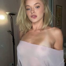 Playful petite blonde independent creator holding her smartphone for a vertical bedroom selfie, wearing a sheer see-through top with visible sun-kissed tan lines.