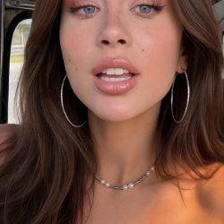 Breathtaking brunette content creator holding her smartphone for a vertical video selfie, smiling and talking casually to the camera under soft bedroom lighting.